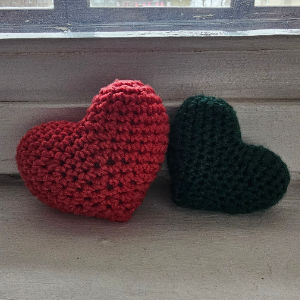 Deux coeurs en crochet, l'un rouge et l'autre vert foncé, reposent côte à côte sur une surface en bois près d'une fenêtre, baignées d'une douce lumière du jour.