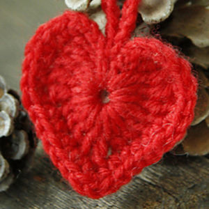 Gros plan d'un ornement en forme de cœur rouge en crochet avec une texture de fil visible, suspendu devant des pommes de pin sur une surface en bois.