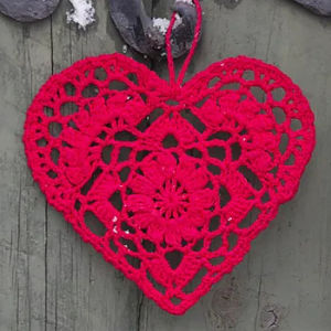 Un ornement en forme de cœur en crochet rouge avec des motifs complexes est suspendu sur un fond en bois patiné.
