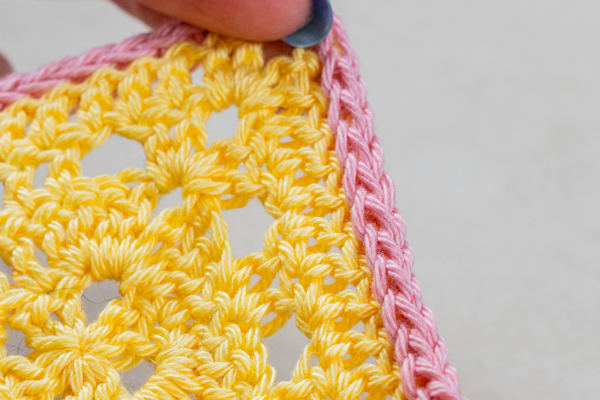 Gros plan d'une main tenant le coin d'une pièce crochetée avec des points jaunes et une bordure rose pâle, mettant en valeur une délicate cordelette au crochet.