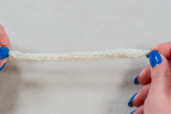 Gros plan de deux mains avec du vernis à ongles bleu tenant une courte chaîne au crochet - également appelée cordelette au crochet - faite de fil blanc sur un fond neutre.
