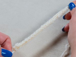 Gros plan de mains aux ongles peints en bleu tenant une cordelette blanche au crochet sur fond de tissu clair.