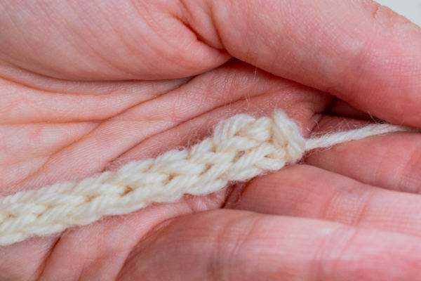 Gros plan sur les mains d'une personne tenant un fil de couleur crème qui a été crocheté pour former une simple cordelette au crochet.