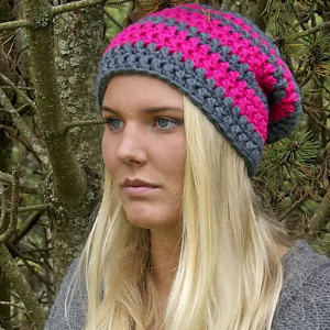 Une femme aux longs cheveux blonds porte un bonnet au crochet à rayures roses et grises, debout à l'extérieur près des branches d'arbres et regardant légèrement sur le côté.