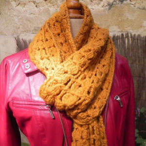 Un mannequin porte une écharpe au crochet texturée jaune moutarde enroulée autour du cou sur une veste en cuir rose vif, posée sur un mur rustique de couleur beige.