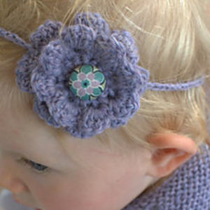 Un jeune enfant aux cheveux blonds portant un bandeau lavande au crochet représentant une grande fleur violette avec un bouton décoratif au centre.