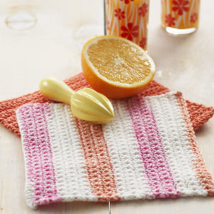 Un torchon à rayures crocheté dans les tons rose, blanc et pêche est posé sur une table à côté d'un torchon orange crocheté, d'un presse-agrumes, d'une orange coupée en deux et de deux verres à motifs floraux.