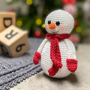 Un bonhomme de neige au crochet avec une écharpe rouge et des mitaines se dresse sur une surface texturée grise, avec des blocs de lettres en bois et un arbre de Noël flou avec des lumières en arrière-plan.