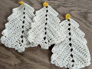 Trois pièces blanches crochetées en forme de sapins de Noël aux cimes jaunes sont disposées sur une surface en bois. La texture du fil et les coutures détaillées en font un accessoire idéal pour les décorations de Noël au crochet.