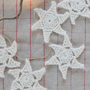 Une guirlande d'étoiles blanches crochetées est disposée sur un fond en tissu à motif quadrillé rouge. Les étoiles sont reliées par une fine ficelle.