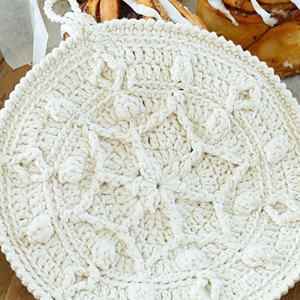 Un manique rond, de couleur crème, crocheté avec un motif texturé et complexe est placé sur une surface en bois près d'une assiette de rouleaux de cannelle.