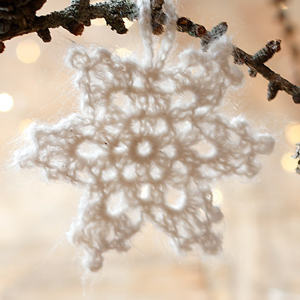 Un flocon de neige blanc crocheté est suspendu à une branche d'arbre, avec des lumières douces et floues qui brillent en arrière-plan.