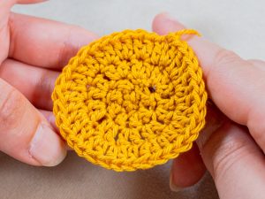 Deux mains tiennent un cercle jaune au crochet à mailles apparentes, probablement réalisé avec du fil selon la technique du crochet en rond, sur un fond neutre.