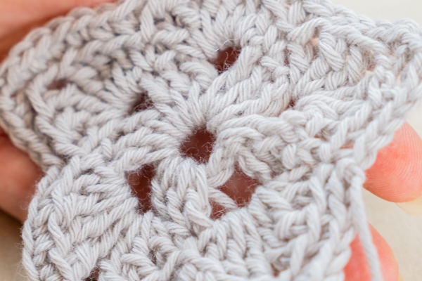 Gros plan d'une main tenant un granny square gris clair au crochet, mettant en valeur le motif de point détaillé, les espaces ouverts et la magnifique technique du crochet double bride au.