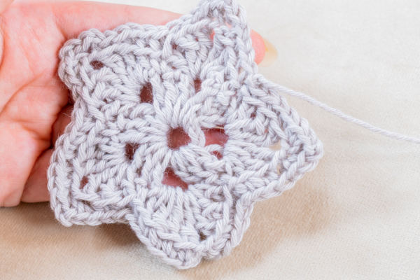 Une main tenant un motif au crochet en forme d'étoile blanche avec un trou au centre, réalisé selon la technique de la double bride au crochet, présenté sur un fond de tissu de couleur claire.