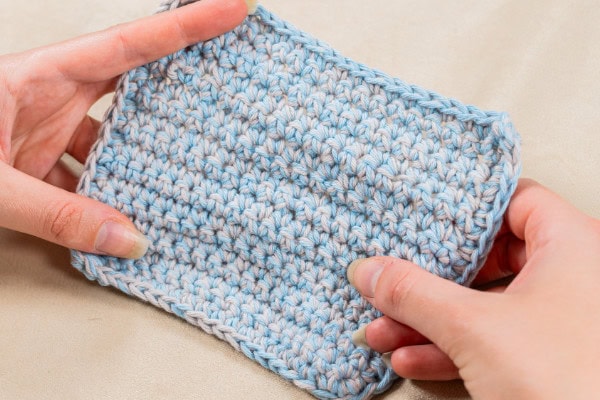 Deux mains tiennent un morceau rectangulaire de tissu crocheté avec du fil panaché bleu clair et blanc, parfait pour tous ceux qui aiment faire du crochet, mettant en valeur la texture et le motif des points sur un fond neutre.