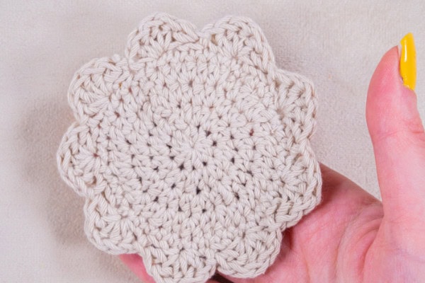 Une main aux ongles peints en jaune tient un sous-verre en forme de fleur crochetée de couleur crème sur un fond clair - une inspiration parfaite pour tous ceux qui aiment faire du crochet.