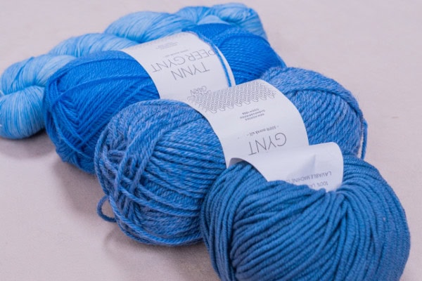 Trois écheveaux de fil bleu de différentes nuances sont empilés sur une surface claire. Chaque écheveau porte une étiquette blanche avec un texte et une marque - parfait pour ceux qui aiment faire du crochet, avec des textures qui varient légèrement en épaisseur et en tonalité de couleur.