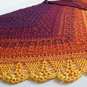 Gros plan d'un châle au crochet avec un motif dégradé, passant du bordeaux foncé en haut au jaune vif en bas, mettant en valeur une dentelle complexe et des points texturés le long du bord.