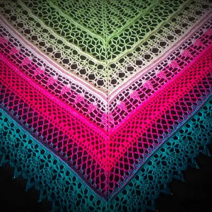 Un châle au crochet triangulaire coloré avec des motifs de dentelle complexes, comportant des rayures dégradées en vert, blanc, rose, magenta et bleu sarcelle, affichées sur un fond sombre.