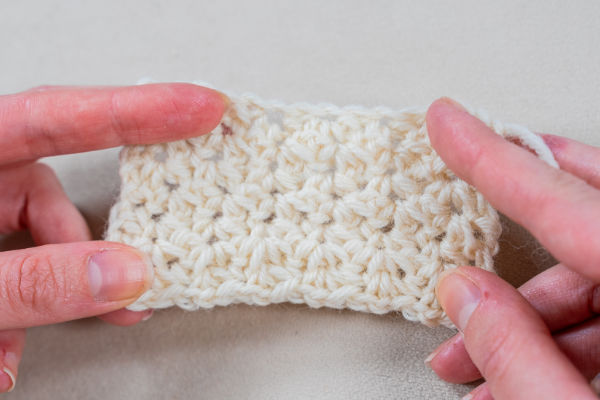 Deux mains tiennent un petit morceau rectangulaire de tissu crocheté blanc cassé, présentant des jolis points au crochet faciles avec un motif de point texturé sur un fond neutre.