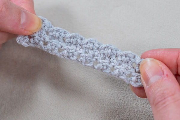 Gros plan de deux mains tenant une courte bande crochetée gris clair, mettant en valeur la texture et les points - parfait pour mettre en valeur des jolis points au crochet faciles sur un fond neutre.