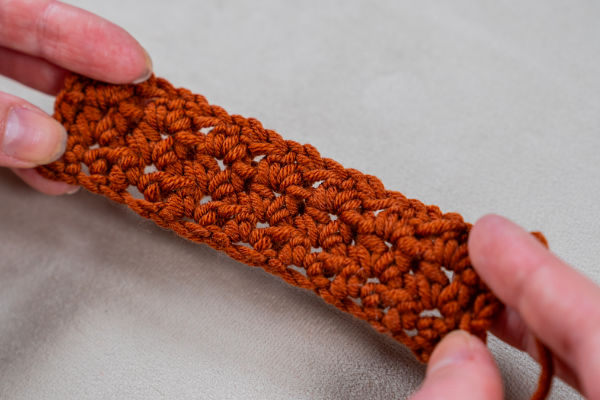 Deux mains tiennent un petit morceau rectangulaire de tissu marron crocheté, mettant en valeur des jolis points au crochet faciles sur un fond doux et clair.