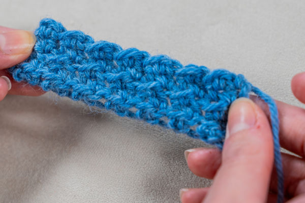 Deux mains tiennent un petit morceau de tissu bleu crocheté, mettant en valeur des jolis points au crochet faciles. Le motif texturé des points se détache sur un fond neutre et clair.