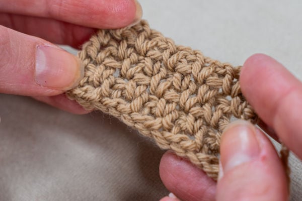 Gros plan de deux mains tenant un petit morceau de tissu au crochet, montrant les détails des jolis points au crochet faciles. L'arrière-plan est une surface unie de couleur claire.