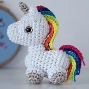 Un charmant jouet licorne au crochet avec un corps blanc, des sabots beiges, une crinière et une queue aux couleurs de l'arc-en-ciel et un grand œil bouton noir, debout sur une surface blanche.