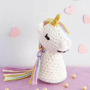 Une licorne au crochet, avec une corne dorée, une crinière colorée et un visage souriant, repose sur une surface violette. Des cœurs roses et de petites perles sont disséminés tout autour.