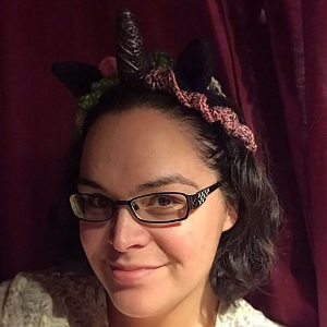 Une personne portant des lunettes et des cheveux noirs mi-longs sourit à la caméra, portant un serre-tête licorne au crochet avec des cornes noires et violettes et une bordure rose, sur fond de rideau rouge foncé.