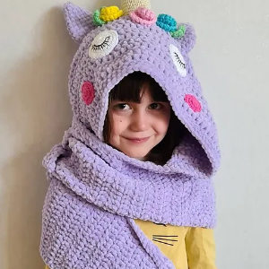 Une jeune fille sourit tout en portant une écharpe à capuche confortable en crochet licorne lavande conçue pour ressembler à une licorne, avec des oreilles, une corne et des décorations colorées.