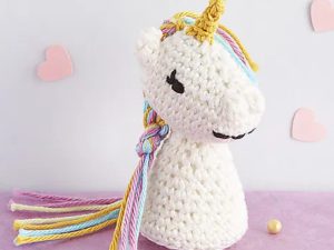 Une charmante licorne au crochet, avec sa corne jaune, sa crinière et sa queue pastel et ses yeux clos, repose sereinement sur une surface rose. Des cœurs roses découpés ornent le mur du fond, ajoutant à son charme fantaisiste.