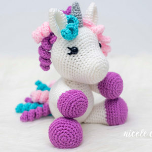 Une charmante peluche licorne au crochet avec un corps blanc, des sabots violets, une crinière et une queue roses et turquoise, des yeux cousus noirs et une corne grise repose sur une surface claire.