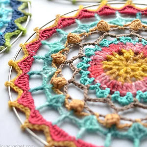 Gros plan d'un mandala crocheté coloré avec des motifs complexes en rose, jaune, bleu sarcelle, bleu et marron, tendu dans un cerceau métallique rond.