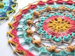 Gros plan d'un motif de mandalas colorés au crochet dans un anneau en métal, comportant des cercles concentriques en fil jaune, rose, bleu et beige avec des points complexes créant un motif décoratif en dentelle.