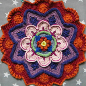 Un mandala crocheté coloré et complexe avec des couches de fils rouges, orange, violets, bleus, verts et roses, affichés sur un fond gris avec des étoiles blanches.
