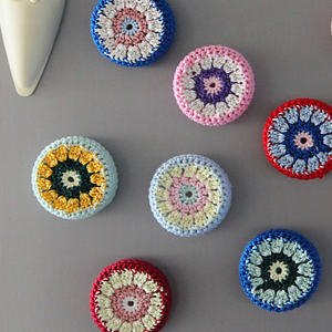 Sept sous-verres ronds et colorés, réalisés au crochet et ornés de divers motifs floraux, sont disposés sur une surface grise. Chaque sous-verre présente des cercles concentriques aux couleurs pastel et vives. Un objet en céramique blanche est partiellement visible dans un coin.
