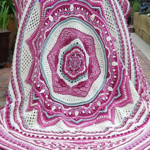 Une grande couverture finement crochetée avec des motifs géométriques concentriques dans des tons de rose, violet, blanc et gris est étalée, montrant sa texture et son design détaillés.