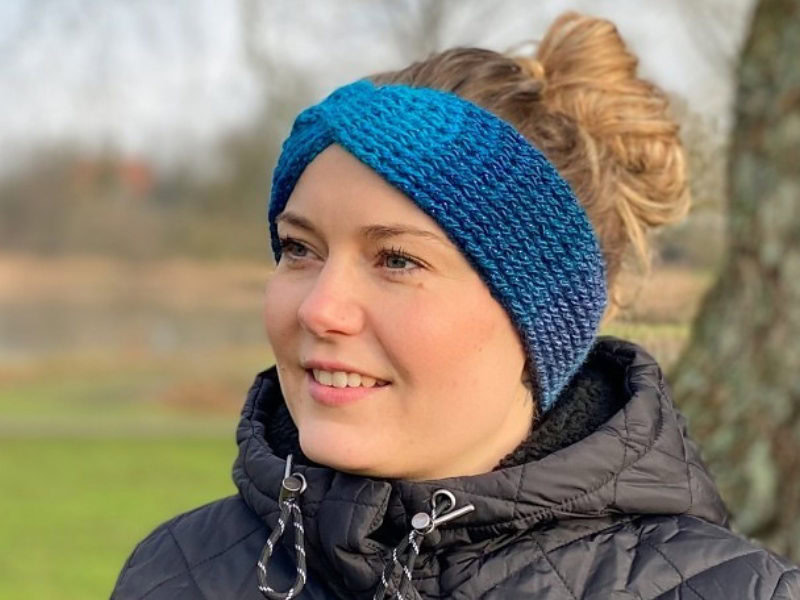 Une femme portant un bandeau bleu tricoté – réalisé à partir d'un modèle de bandeau au crochet gratuit – et une veste matelassée noire se tient dehors, souriante, les cheveux relevés en chignon. L'arrière-plan flou présente des arbres et de l'herbe.