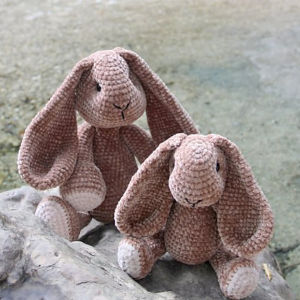 Deux lapins bruns crochetés à la main, aux longues oreilles tombantes, sont assis sur un rocher, face à l'avant. Une source d'inspiration idéale pour tous ceux qui recherchent un modèle gratuit de doudous au crochet. Le fond présente un fond texturé.