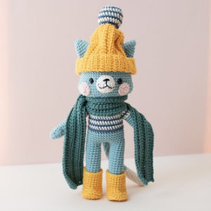Une poupée chat bleu crochetée, réalisée à partir d'un modèle gratuit de doudous au crochet, porte un bonnet en tricot jaune avec un pompon, une écharpe verte, des bottes jaunes et un pull rayé, debout sur un fond rose clair.