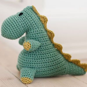 Une peluche dinosaure verte crochetée avec des pieds jaunes et une crête jaune le long du dos, posée sur un sol en bois clair — une inspiration parfaite pour votre prochain modèle gratuit de doudous au crochet.