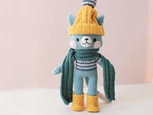 Une poupée chat au crochet — modèle gratuit de doudous au crochet — porte un chapeau jaune avec un pompon, une chemise rayée bleue et blanche, une écharpe verte et des bottes jaunes, se tenant sur un fond rose clair.