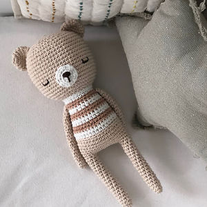 Un ours en peluche au crochet avec les yeux fermés, un museau blanc et un corps rayé beige et blanc repose sur une surface de couleur claire à côté d'un coussin gris, parfait comme modèle gratuit de doudous au crochet.