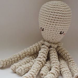 Une pieuvre crochetée à la main avec une tête ronde, des yeux noirs simples, des joues roses et huit tentacules bouclés, fabriquée à partir de fil de couleur crème et inspirée d'un modèle gratuit de doudous au crochet.