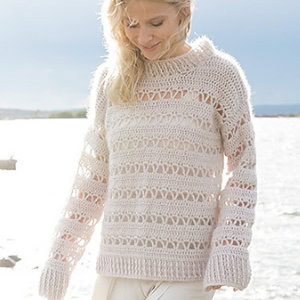 Une femme aux cheveux blonds se tient au bord de l'eau, vêtue d'un pull rose clair, ample, à manches longues, en crochet, orné de motifs ajourés en dentelle. Elle baisse les yeux et sourit doucement.