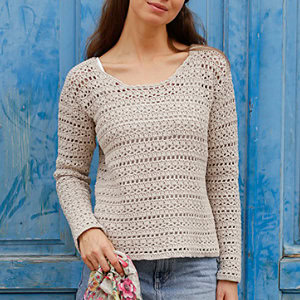 Une femme portant un pull beige au crochet et un jean bleu se tient devant une porte en bois bleu vif, tenant un tissu à motifs floraux dans une main.