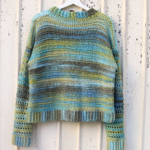 Un pull crocheté multicolore à manches longues est accroché à un mur en bois blanc. Il présente des rayures horizontales dans des tons de bleu, vert, marron et jaune.